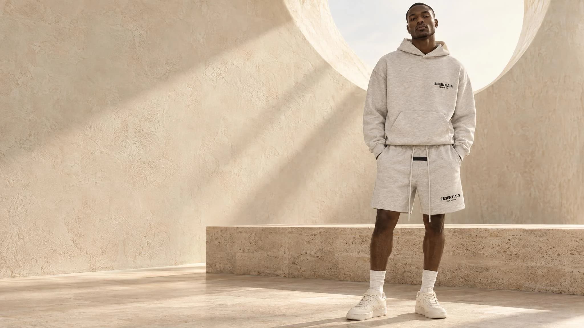 Yenoto AI campaign: a model wearing a light oatmeal Essentials hoodie and sweat shorts beneath a circular window
